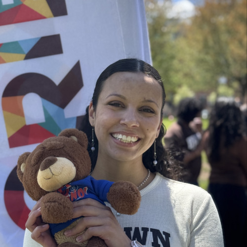 Berenika smiling and posing with a Bruno stuffy outside in front of a New Student Orientation flag