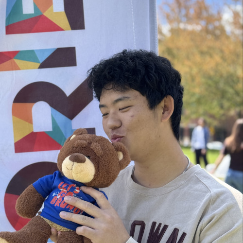 Brady smiling and posing with a Bruno stuffy outside in front of a New Student Orientation flag