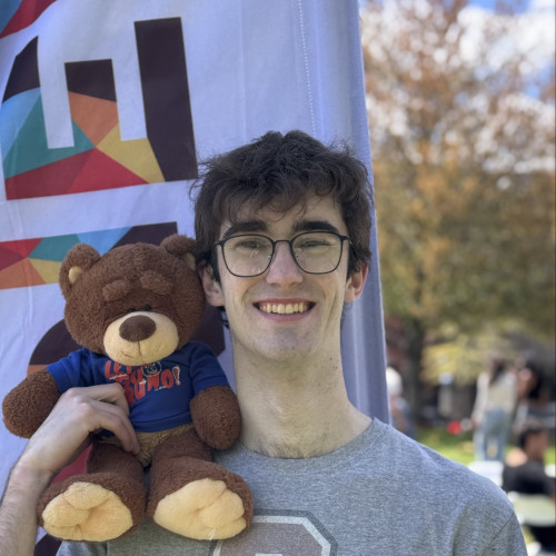 Charles smiling and posing with a Bruno stuffy outside in front of a New Student Orientation flag