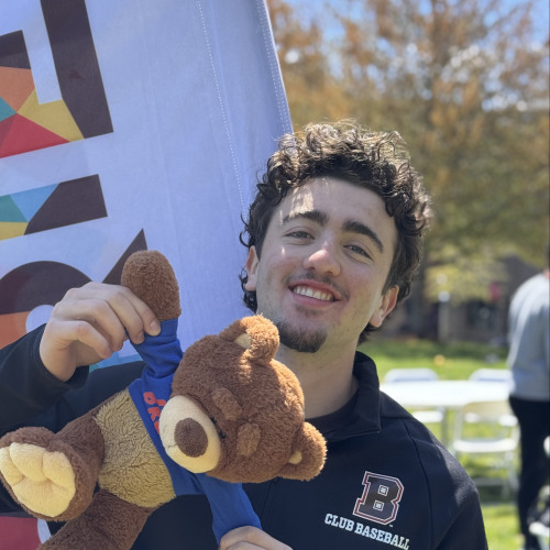 Charlie smiling and posing with a Bruno stuffy outside in front of a New Student Orientation flag