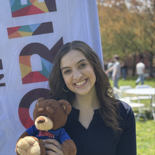 Ella smiling and posing with a Bruno stuffy outside in front of a New Student Orientation flag