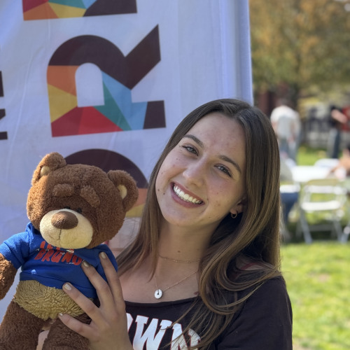 Emma smiling and posing with a Bruno stuffy outside in front of a New Student Orientation flag
