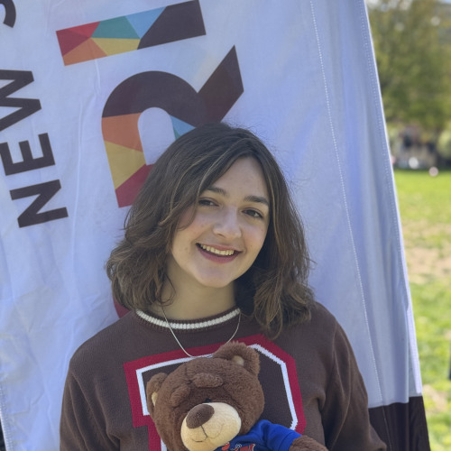 Gaby smiling and posing with a Bruno stuffy outside in front of a New Student Orientation flag