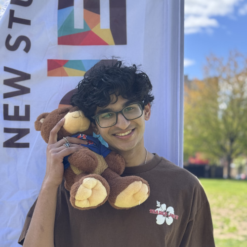 Ishaan smiling and posing with a Bruno stuffy outside in front of a New Student Orientation flag