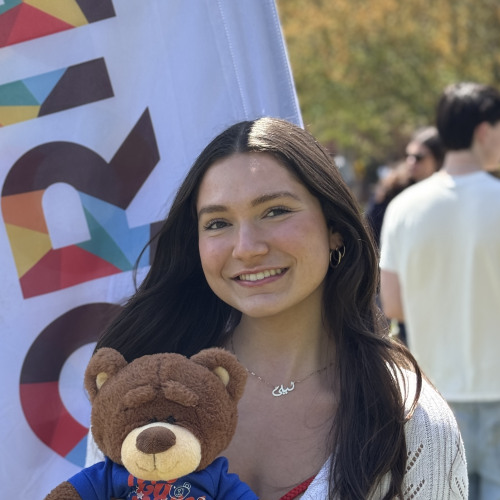 Leila posing with a bruno stuffy and smiling outside in front of an orientation flag