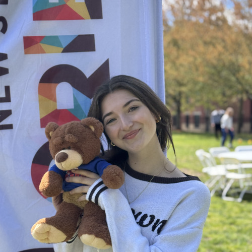 Maya posing with a bruno stuffy and smiling outside in front of an orientation flag