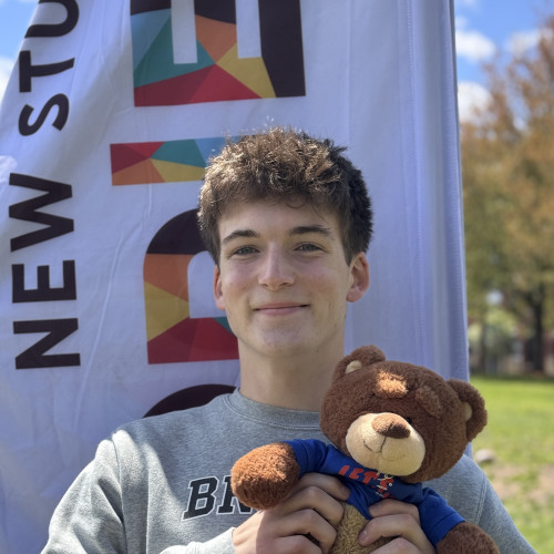 Miles posing with a bruno stuffy and smiling outside in front of an orientation flag