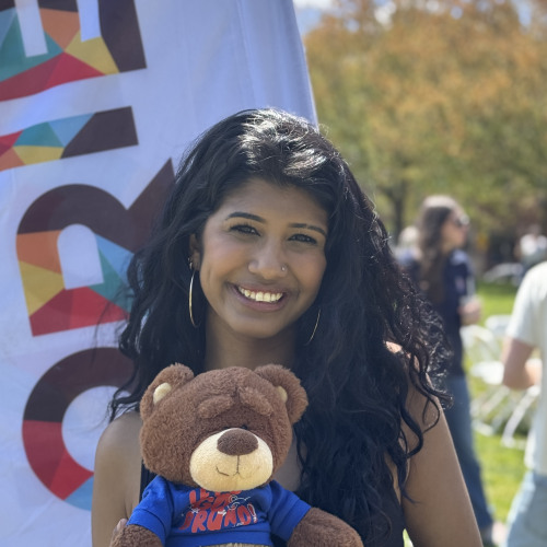 Ramya posing with a bruno stuffy and smiling outside in front of an orientation flag