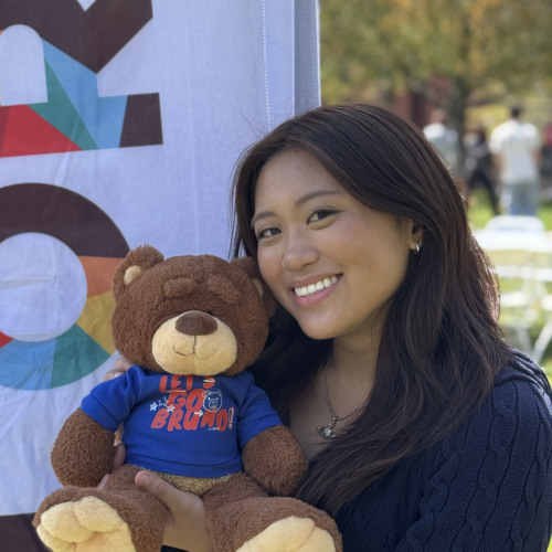 Zoe posing with a bruno stuffy and smiling outside in front of an orientation flag