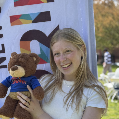 Annika smiling and posing with a Bruno stuffy outside in front of a New Student Orientation flag