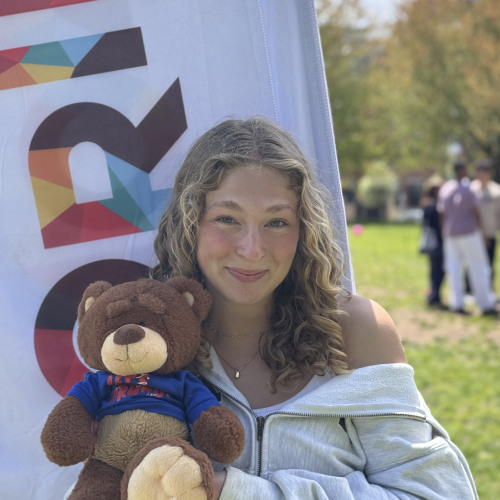 Hannah smiling and posing with a Bruno stuffy outside in front of a New Student Orientation flag