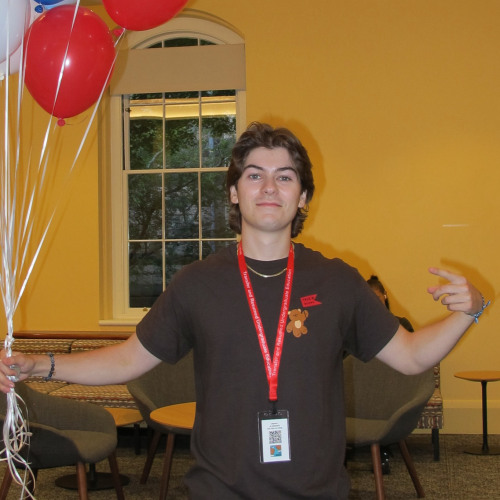 Chip wearing a Brown TRUE Shirt holding a collection of light blue, dark blue, red, and white balloons 